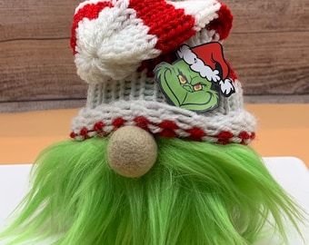 Peut inclure: Un gnome vert portant un chapeau rayé blanc et rouge. Le chapeau a un patch de visage du Grinch sur le devant.