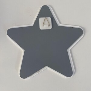 May include: A gray star-shaped wall decoration with a white border. A metal hanger is attached to the back for easy mounting. The star is a solid color and has a clean, modern design. Perfect for a nursery or child's room.