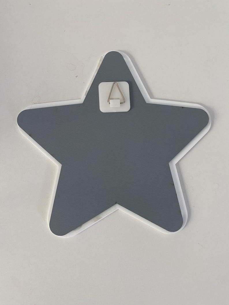 May include: A gray star-shaped wall decoration with a white border. A metal hanger is attached to the back for easy mounting. The star is a solid color and has a clean, modern design. Perfect for a nursery or child's room.