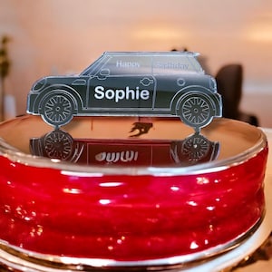 Puede incluir: Una silueta en blanco y negro de un coche mini con las palabras "Happy Birthday Sophie" en el lateral. El coche está encima de un pastel rojo.