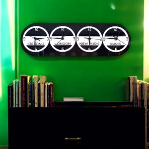 Puede incluir: Un reloj mundial en blanco y negro con cuatro zonas horarias: Irlanda, Londres, Nueva York y París. El reloj está montado en una pared verde sobre un estante negro con libros.