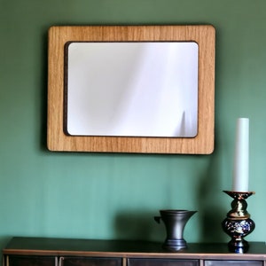 May include: A wooden framed mirror with a light brown wood frame and a white mirror surface. The mirror is hanging on a green wall.