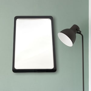 Op de afbeelding: Een rechthoekige spiegel met een zwarte lijst en een witte achtergrond hangt aan een lichtgroene muur. Rechts van de spiegel staat een zwarte lamp met een witte kap.