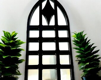 May include: Black gothic-style mirror with a diamond-shaped accent at the top. The mirror is framed with a grid pattern and is flanked by two small potted green plants. The mirror is a decorative home decor item.