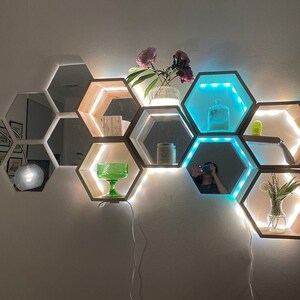 Könnte beinhalten: Ein Satz hexagonaler Regale mit verspiegelten Fronten und LED-Leuchten. Die Regale sind in einem Wabenmuster angeordnet und mit blauen und weißen Lichtern beleuchtet. Die Regale sind mit verschiedenen Gegenständen dekoriert, darunter eine Glasvase mit Blumen, eine Glasschüssel und eine Kerze.