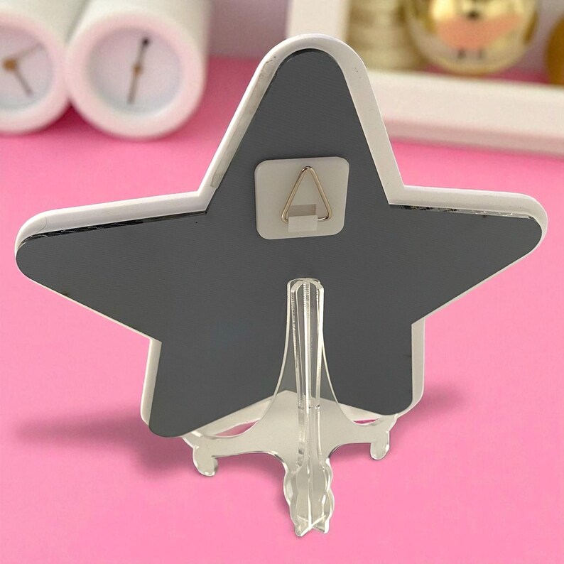 May include: A star-shaped decorative item with a gray center and white trim. It has a clear plastic stand and a small hook on the back for hanging. The item is displayed on a pink surface.