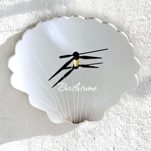 Peut inclure: Une horloge murale en miroir en forme de coquille avec des aiguilles noires et le mot "Bathtime" imprimé sur le cadran.