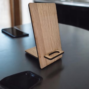 May include: A wooden phone stand with a natural wood grain finish. The stand has a vertical back and a small shelf at the bottom to hold a phone. The stand is designed to hold a phone upright for hands-free use.