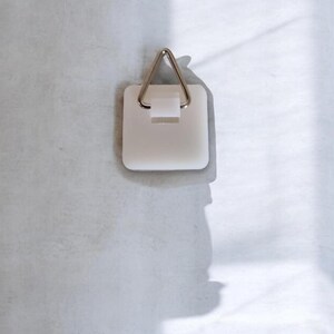 May include: A white, square picture hanger with a metal triangle hook. The hanger is designed to be mounted on a wall for hanging pictures or other decorative items. The hook is silver in color.