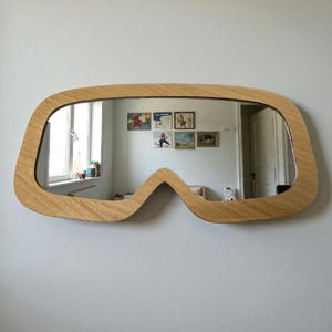 May include: A distinctive wall mirror with a light brown wooden frame. The mirror's shape is unconventional, reminiscent of goggles or a stylized face. The reflection reveals a room with a window and artwork.