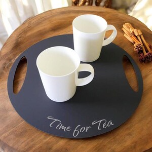 Puede incluir: Una bandeja redonda de servir de color gris oscuro con dos tazas de cerámica blanca y asas a cada lado. La bandeja tiene las palabras "Time for Tea" escritas en ella con una fuente cursiva.