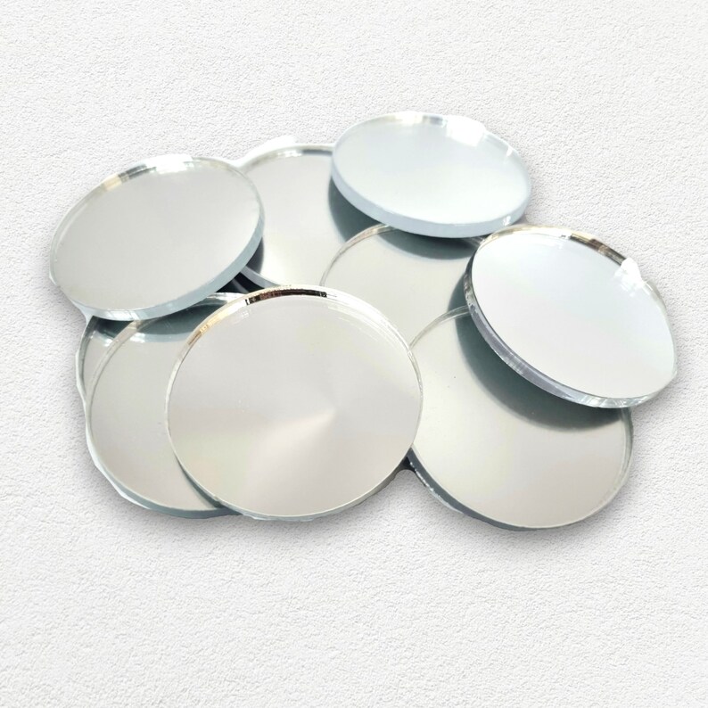 May include: A pile of round, silver-colored, reflective mirror tiles. The tiles are arranged in a random pattern, with some overlapping.
