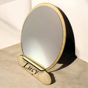 Puede incluir: Un espejo redondo de madera con un marco de madera clara y un reflejo claro. El espejo está de pie sobre una base de madera con el nombre "Lucy" grabado en él.