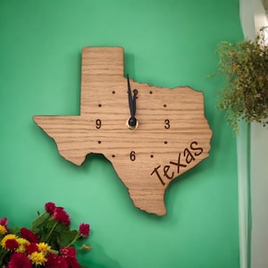May include: A wooden clock shaped like the state of Texas with the word "Texas" inscribed on the bottom. The clock face has black hands and Roman numeral markings.