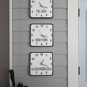 Puede incluir: Tres relojes cuadrados blancos con números y manecillas negros, montados en una pared gris. Los relojes muestran la hora en Tel Aviv, Nueva York y Los Ángeles.
