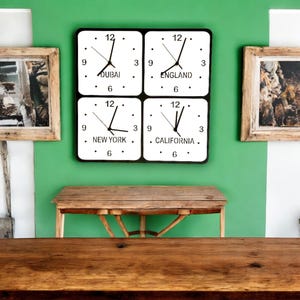 Puede incluir: Un conjunto de cuatro relojes con marcos negros y caras blancas, cada uno mostrando una zona horaria diferente: Dubái, Inglaterra, Nueva York y California. Los relojes están montados en una pared verde con una mesa de madera en primer plano.