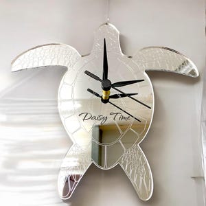 Könnte beinhalten: Eine verspiegelte Wanduhr in Form einer Meeresschildkröte. Die Uhr hat schwarze Zeiger und die Worte "Daisy Time" in einer Schreibschrift. Sie hängt vor einem weißen Hintergrund.