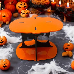 Puede incluir: Soporte para pastel en forma de calabaza naranja y negro con base negra y cuatro patas. El soporte está decorado con formas de calabaza recortadas.