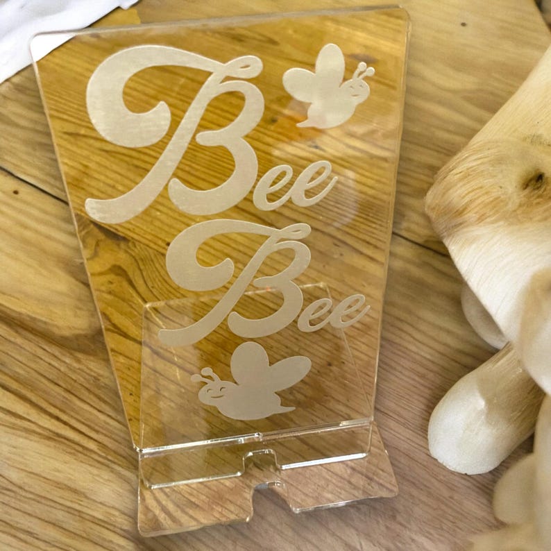 May include: Clear acrylic phone stand with the word "Bee" in a cursive font, along with two bee illustrations. The stand has a slot at the bottom for a phone and is on a wooden surface.