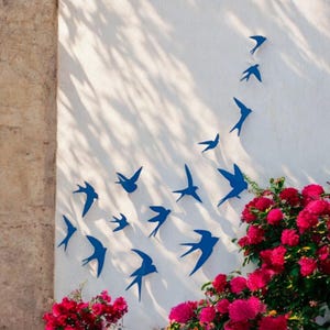 Puede incluir: Una pared blanca con un grupo de siluetas de pájaros azules, dispuestas en un patrón fluido. Los pájaros están todos mirando en la misma dirección, creando una sensación de movimiento. Se ven flores rosas en las esquinas inferiores de la imagen.