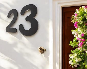 May include: Black house number 23 with a white background. The number is attached to a white wall next to a wooden door with a floral wreath.