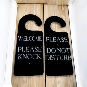 Pode incluir: Duas placas de porta pretas com a parte superior curva. A placa esquerda diz "WELCOME PLEASE KNOCK" em texto branco. A placa direita diz "PLEASE DO NOT DISTURB" em texto branco. As placas estão montadas numa porta de madeira clara.