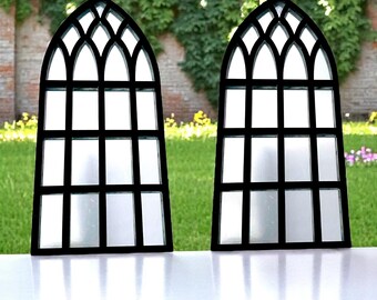 May include: Two black metal framed windows with arched tops and frosted glass panes. The windows are set against a backdrop of green foliage and a brick wall.