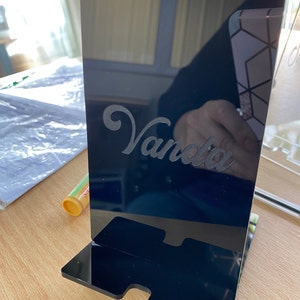 May include: A black acrylic phone stand with the name "Vanda" etched in a cursive font. The stand has a rectangular base with a slot for the phone and a vertical panel. The stand is on a wooden surface.