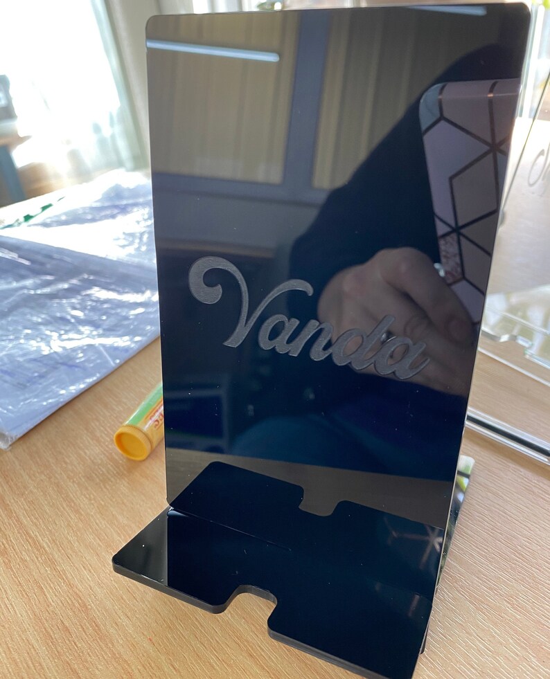 May include: A black acrylic phone stand with the name "Vanda" etched in a cursive font. The stand has a rectangular base with a slot for the phone and a vertical panel. The stand is on a wooden surface.