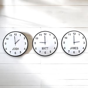 May include: Three round wall clocks with black rims and white faces are displayed. Each clock has black numbers and hands. The first clock reads "JOSH" at the bottom, the second reads "MATT", and the third reads "JAMES".