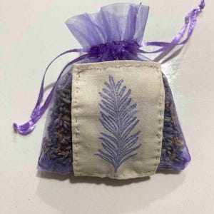 Puede incluir: Una pequeña bolsita llena de lavanda con un cierre de cordón morado transparente. La bolsita tiene una etiqueta rectangular de tela blanquecina con un diseño de helecho morado. La bolsita está llena de capullos de lavanda secos.