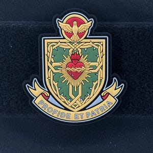 May include: A patch featuring a shield design with a red heart, a gold cross, and a dove at the top. The patch has the text "PROFIDE ET PATRIA" on a banner. The patch is set against a black background.