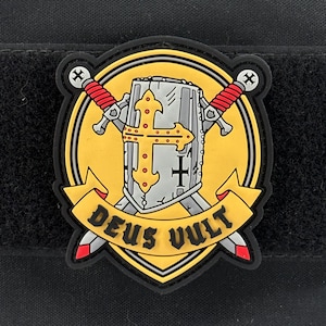 May include: A patch featuring a yellow and black design with a knight's helmet, a cross, and two crossed swords with red handles. The words "DEUS VULT" are on a yellow banner.