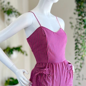 May include: A pink and black striped jumpsuit with a sweetheart neckline and two pockets on the front. The jumpsuit has thin straps and a fitted bodice.