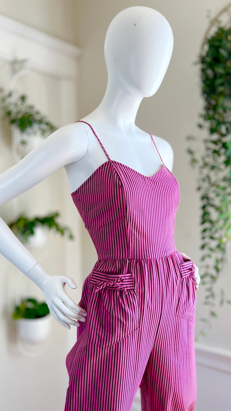 May include: A pink and black striped jumpsuit with a sweetheart neckline and two pockets on the front. The jumpsuit has thin straps and a fitted bodice.