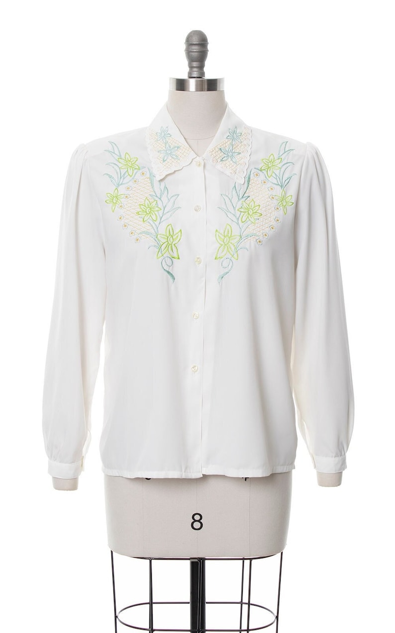 Puede incluir: Camisa blanca de manga larga con botones, cuello de encaje y bordado floral.