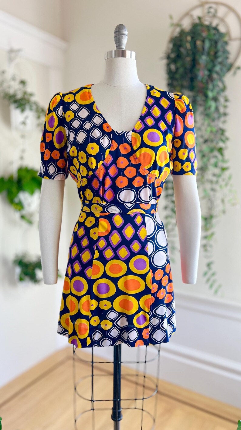 May include: A vintage dress with a bold geometric design. The dress has a V-neck, short sleeves, and a fitted waist. The pattern incorporates orange, yellow, purple, and white shapes on a navy blue backdrop. The dress is shown on a mannequin.