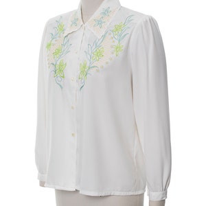 Puede incluir: Una blusa blanca de manga larga con cierre de botones y un dise&ntilde;o bordado de flores en el cuello y el pecho.
