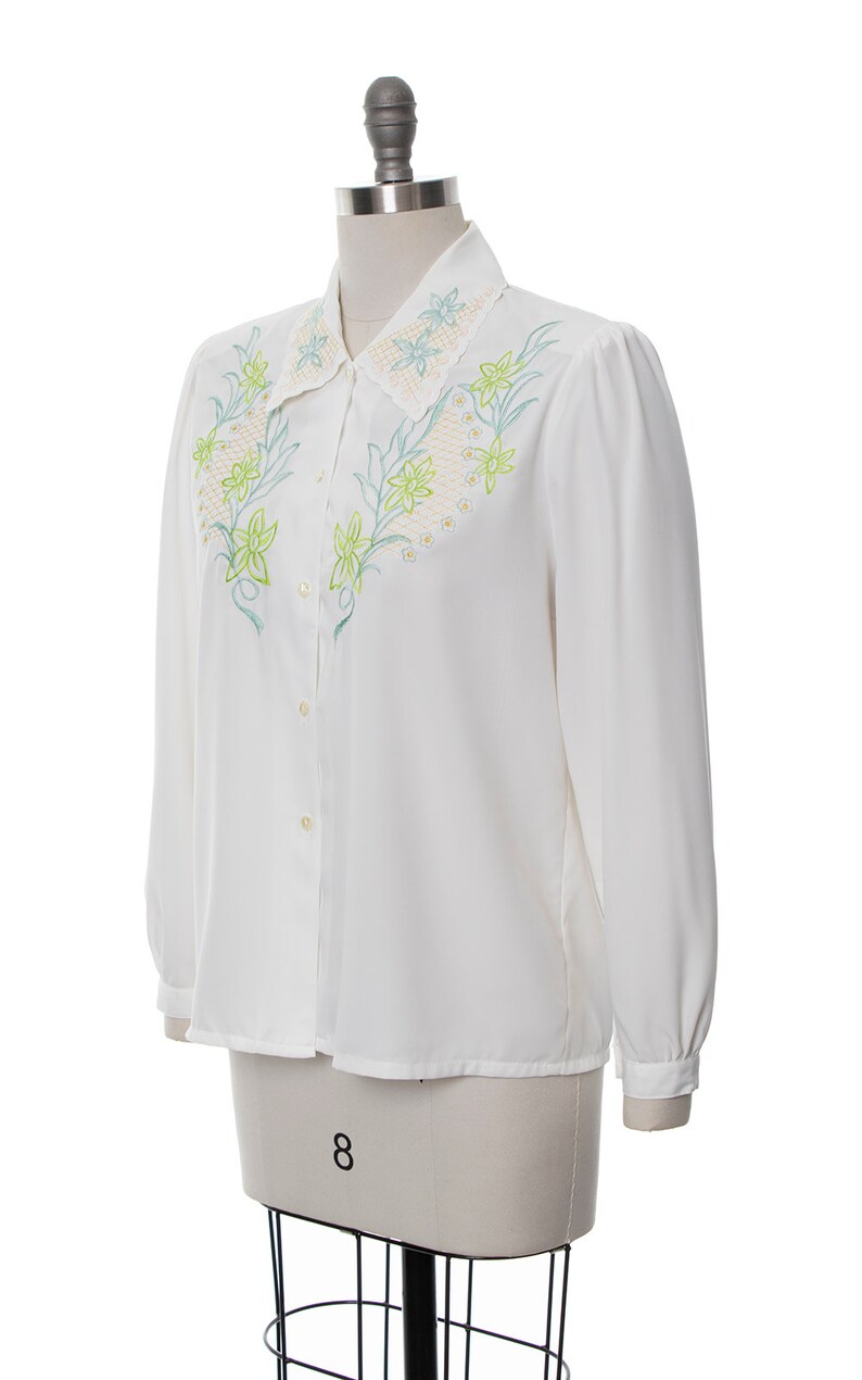 Puede incluir: Una blusa blanca de manga larga con cierre de botones y un dise&ntilde;o bordado de flores en el cuello y el pecho.