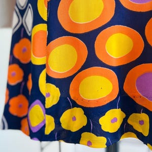 May include: Navy blue fabric with a striking pattern of orange, yellow, and purple geometric shapes and floral designs. The fabric features large circles and smaller flower shapes, creating a bold and colourful textile.