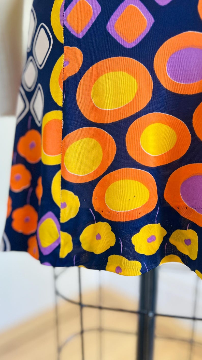 May include: Navy blue fabric with a striking pattern of orange, yellow, and purple geometric shapes and floral designs. The fabric features large circles and smaller flower shapes, creating a bold and colourful textile.