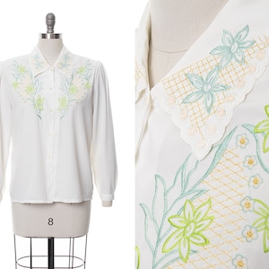 Puede incluir: Una blusa blanca de manga larga con un delicado dise&ntilde;o de bordado floral en tonos verdes y amarillos. El cuello est&aacute; adornado con un borde festoneado y un intrincado bordado floral.
