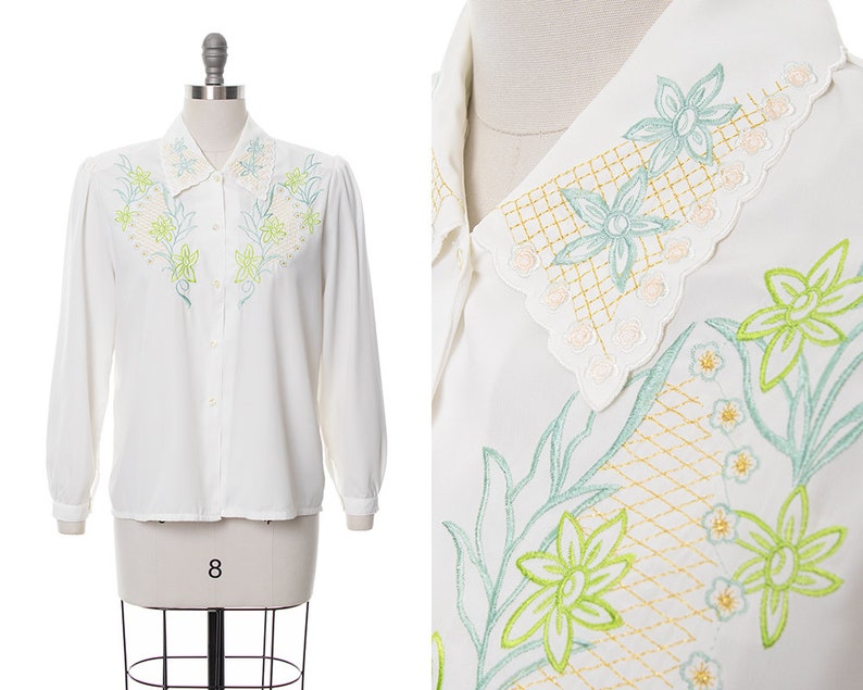 Puede incluir: Una blusa blanca de manga larga con un delicado dise&ntilde;o de bordado floral en tonos verdes y amarillos. El cuello est&aacute; adornado con un borde festoneado y un intrincado bordado floral.