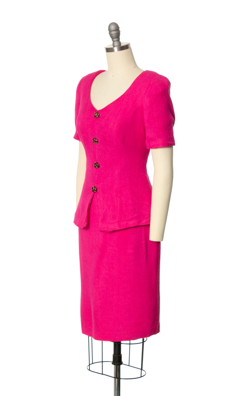 May include: A pink short-sleeved dress suit with a skirt and a fitted jacket. The jacket has a V-neckline and a button-up front.