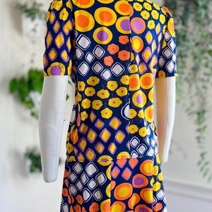 May include: A retro-style dress with a striking pattern of orange, yellow, purple, and white shapes on a navy blue background. The dress has short sleeves and a knee-length hemline, displayed on a mannequin.