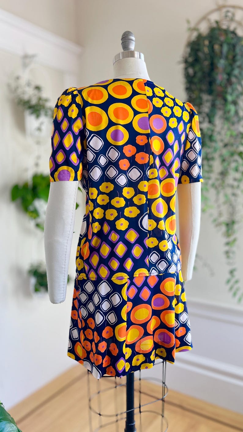 May include: A retro-style dress with a striking pattern of orange, yellow, purple, and white shapes on a navy blue background. The dress has short sleeves and a knee-length hemline, displayed on a mannequin.