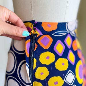 May include: A patterned skirt with a navy blue base, adorned with yellow flowers, orange and purple geometric shapes, and white circles. The skirt's gold zipper is partially open. The design is eye-catching and bold.