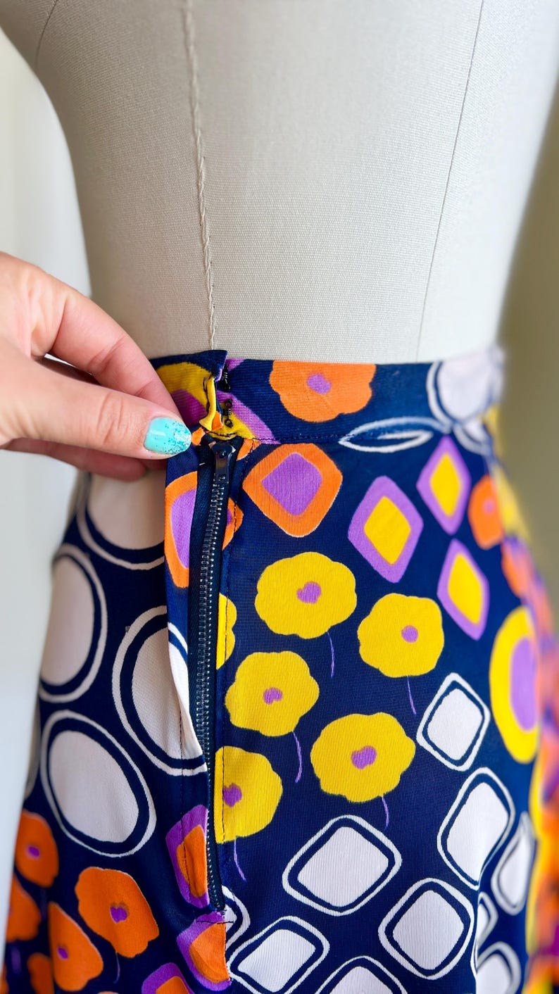 May include: A patterned skirt with a navy blue base, adorned with yellow flowers, orange and purple geometric shapes, and white circles. The skirt's gold zipper is partially open. The design is eye-catching and bold.