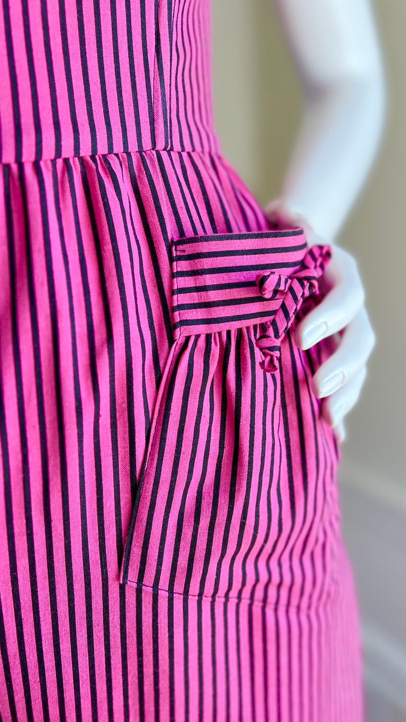 May include: A pink and black striped dress with a patch pocket on the side. The pocket has a bow detail.