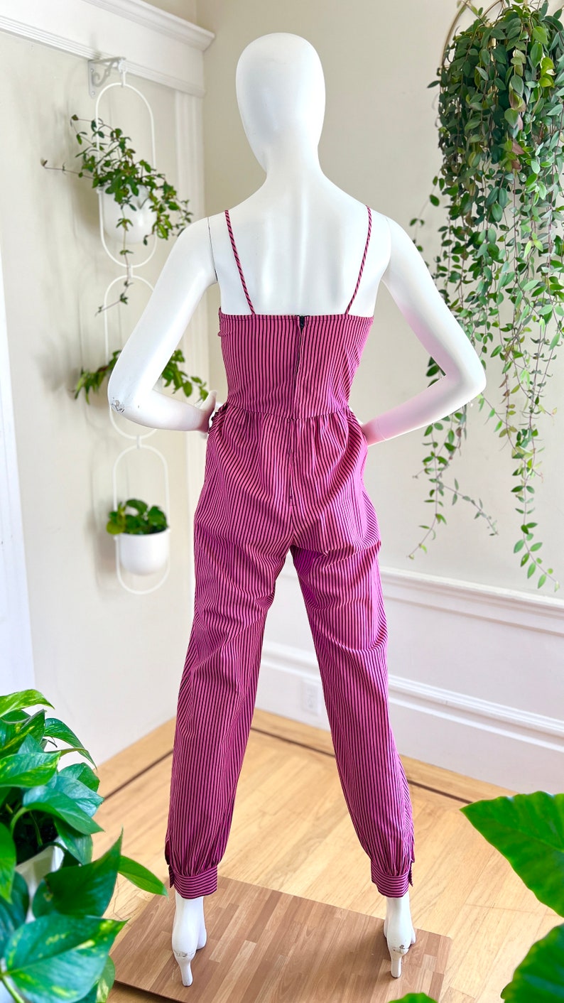 May include: A pink and black striped jumpsuit with thin straps and a fitted bodice. The jumpsuit has a relaxed fit through the legs and features a cuffed hem.